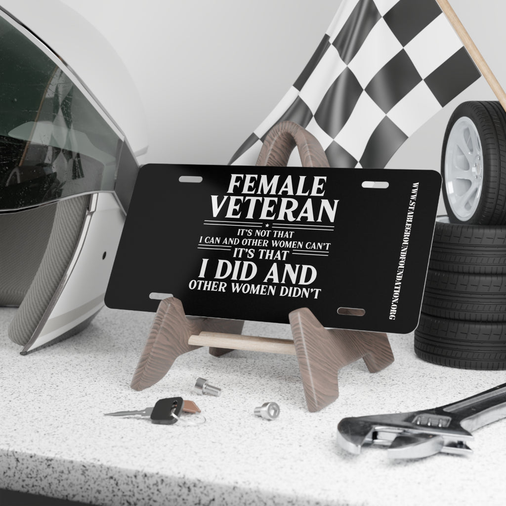 Black license plate with bold white text reading: “Female Veteran. It’s not that I can and other women can’t, it’s that I did and other women didn’t.” Displayed on a stand with tools, keys, helmet, and checkered flag in the background.