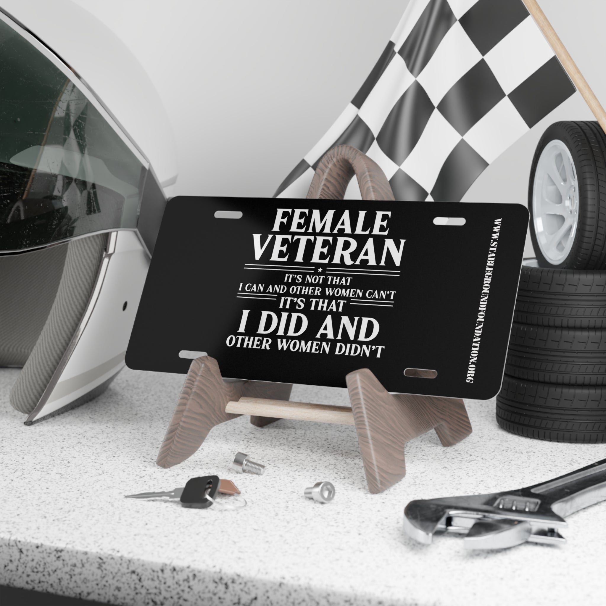 Black license plate with bold white text reading: “Female Veteran. It’s not that I can and other women can’t, it’s that I did and other women didn’t.” Displayed on a stand with tools, keys, helmet, and checkered flag in the background.