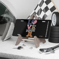 Black license plate with the word “LOVE” in bold patriotic letters: American flag heart for “O,” camo “V,” combat boots as “E,” and text reading “Our Veterans.” Displayed on a stand with tools, keys, and racing gear in the background.