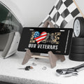 Black license plate with the word “LOVE” in camo letters, an American flag heart, and tan combat boots. Text below reads “Our Veterans.” Displayed on a stand with racing gear and tools.