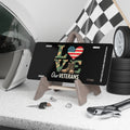 Black license plate with the word “LOVE” in camouflage letters, an American flag heart, and tan combat boots. Text below reads “Our Veterans.” Displayed on a stand with racing gear and tools.