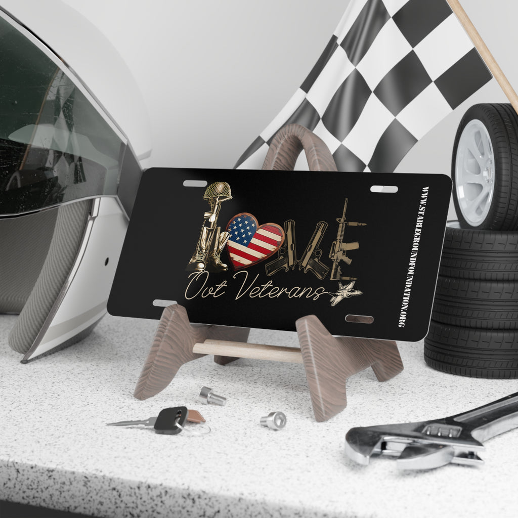 Black license plate featuring the word “LOVE” spelled out using a battlefield cross helmet and rifle, an American flag heart, and military rifles. Text below reads “Our Veterans.” Displayed on a stand with racing gear and tools.