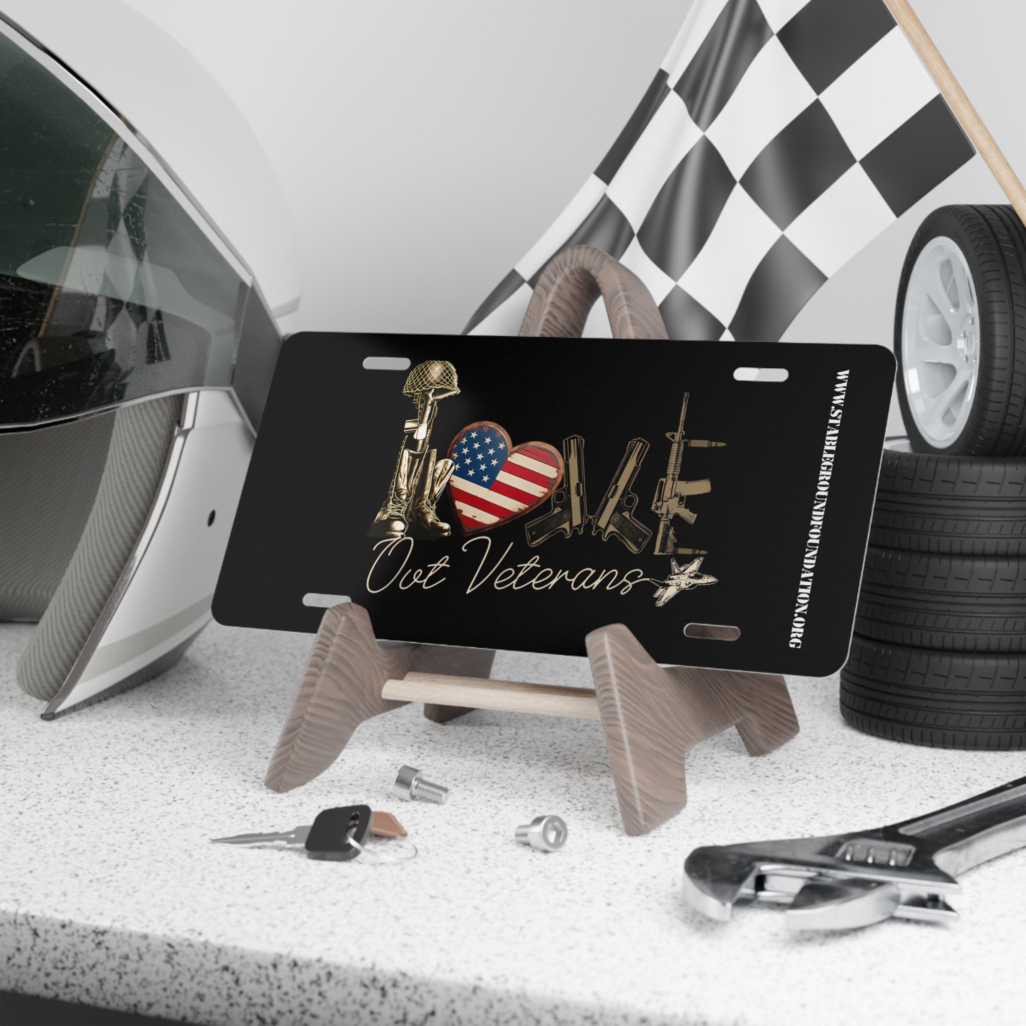 Black license plate featuring the word “LOVE” spelled out using a battlefield cross helmet and rifle, an American flag heart, and military rifles. Text below reads “Our Veterans.” Displayed on a stand with racing gear and tools.