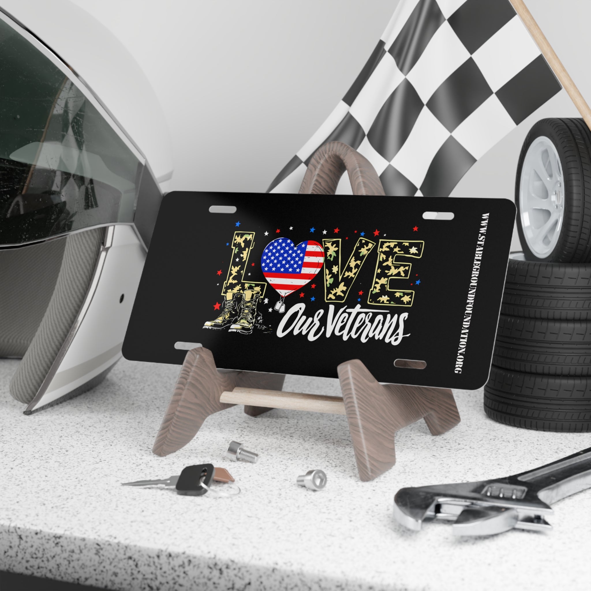 Black license plate with the word “LOVE” in camouflage letters filled with yellow stars, an American flag heart, and combat boots. Below text reads “Our Veterans.” Displayed on a stand with racing gear and tools in the background.