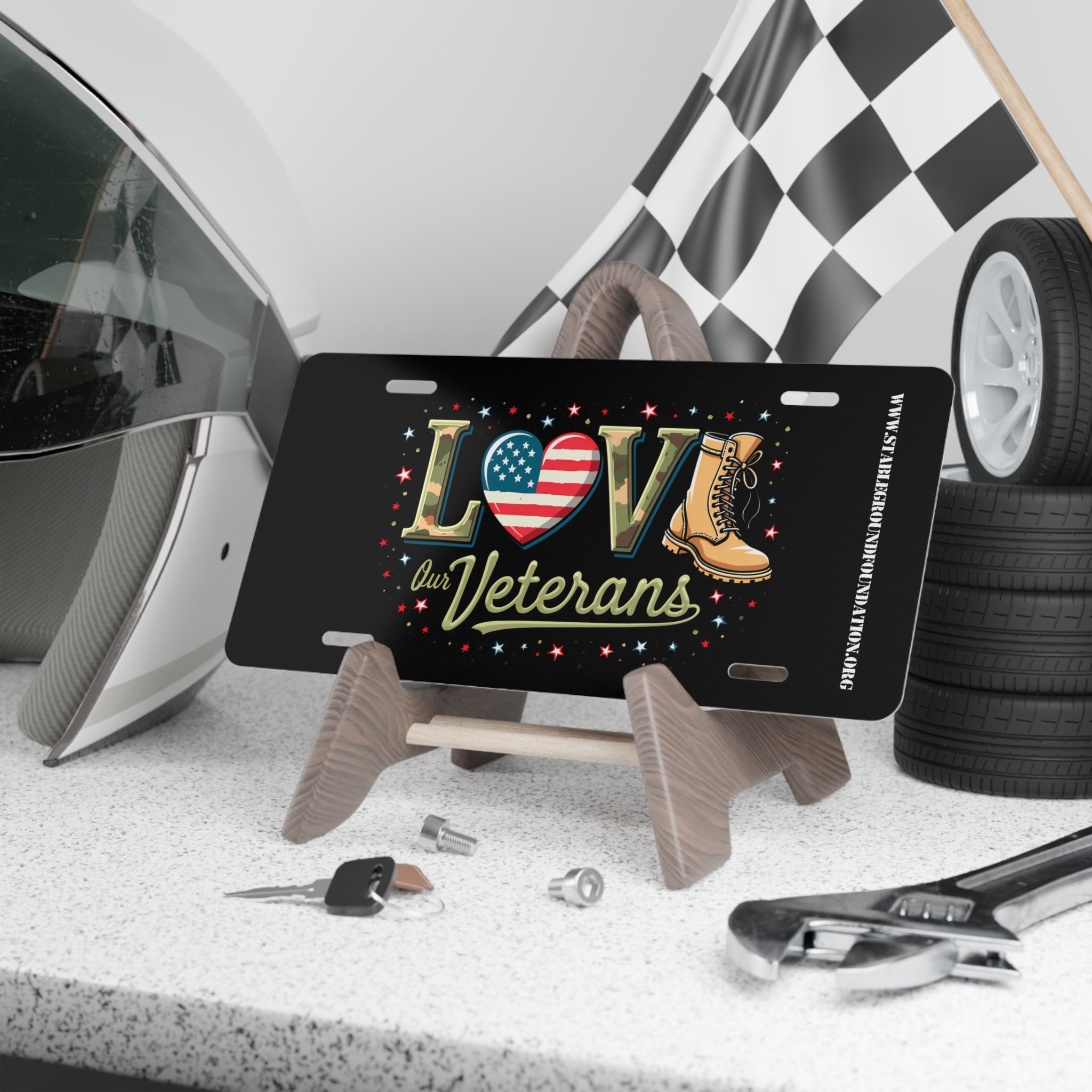 Black license plate with the word “LOVE” in bold letters: American flag heart for “O,” camo-style lettering, and tan combat boots as part of the design. Surrounded by red, white, and blue stars with the words “Our Veterans.” Displayed on a stand with racing gear and tools in the background.