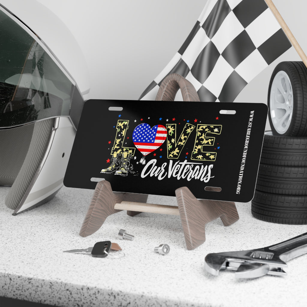 Black license plate with the word “LOVE” in camouflage letters decorated with stars, an American flag heart, and combat boots. Text below reads “Our Veterans.” Displayed on a stand with racing gear and tools.