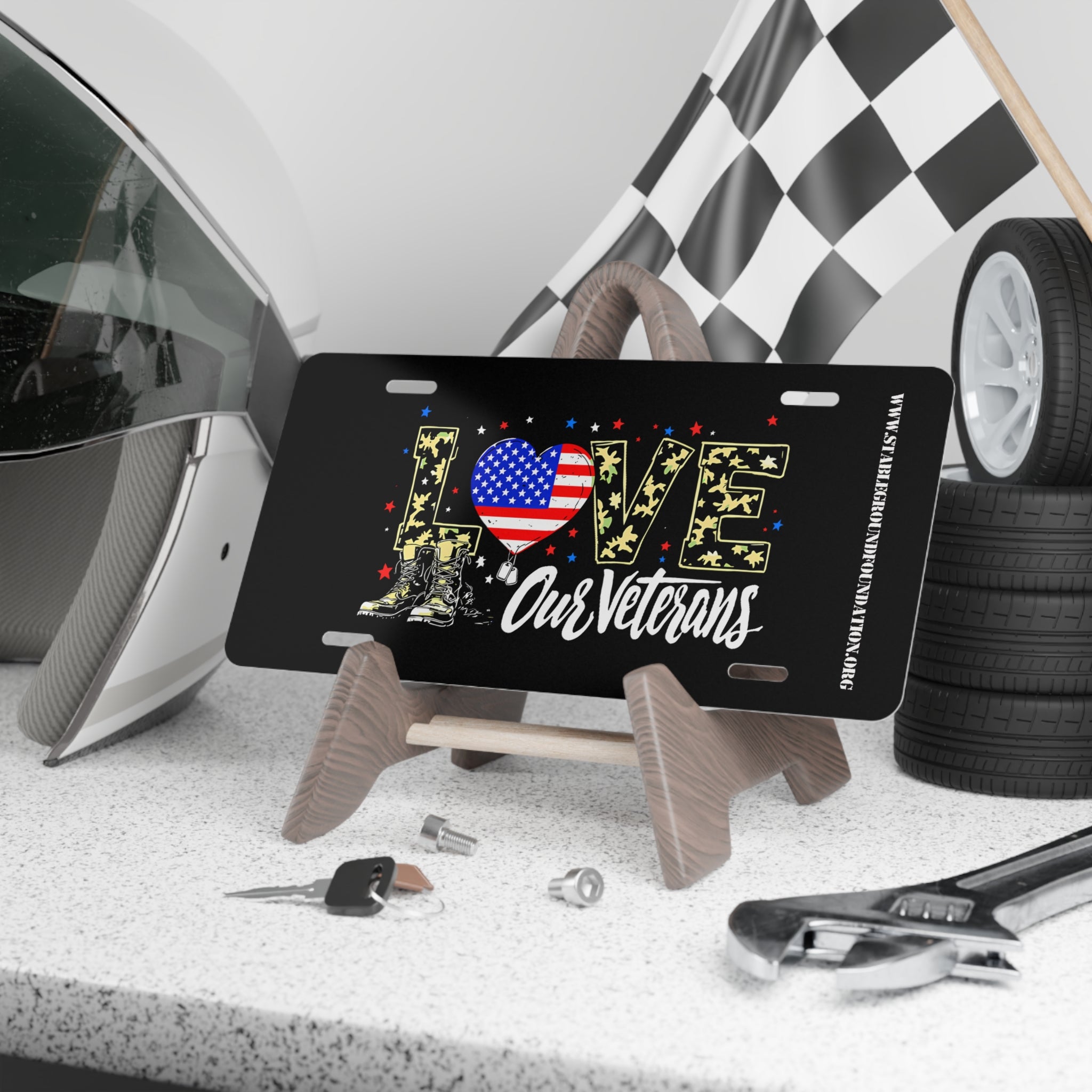 Black license plate with the word “LOVE” in camouflage letters decorated with stars, an American flag heart, and combat boots. Text below reads “Our Veterans.” Displayed on a stand with racing gear and tools.
