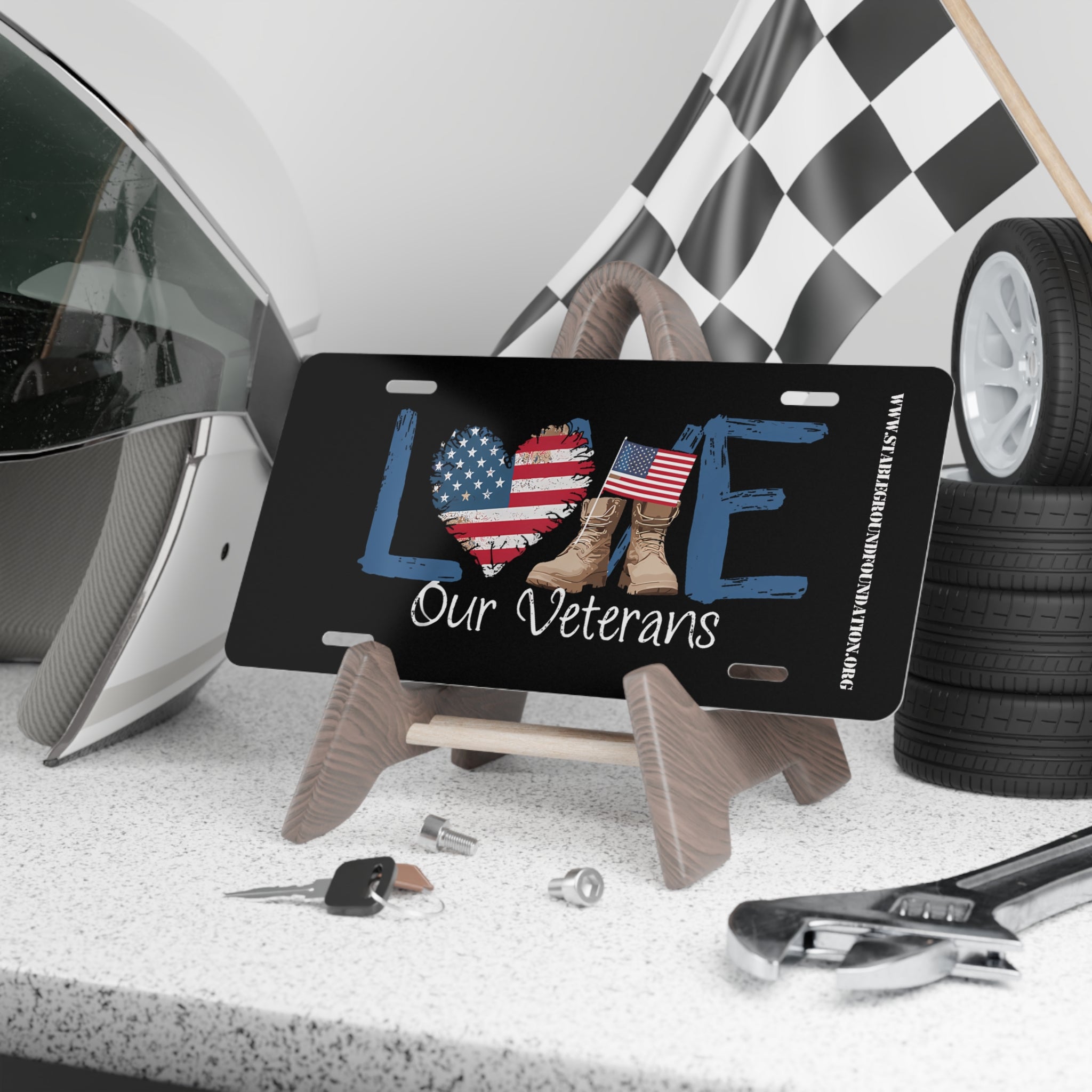Black license plate with the word “LOVE” in distressed blue letters, American flag heart, tan combat boots, and text reading “Our Veterans.” Displayed on a wooden stand with helmet, checkered flag, tools, and keys in the background.