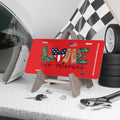Red license plate with the word “LOVE” spelled out using camouflage letters, an American flag heart, tan combat boots, and a small waving U.S. flag. Text below reads “Our Veterans.” Displayed on a stand with racing gear and tools.