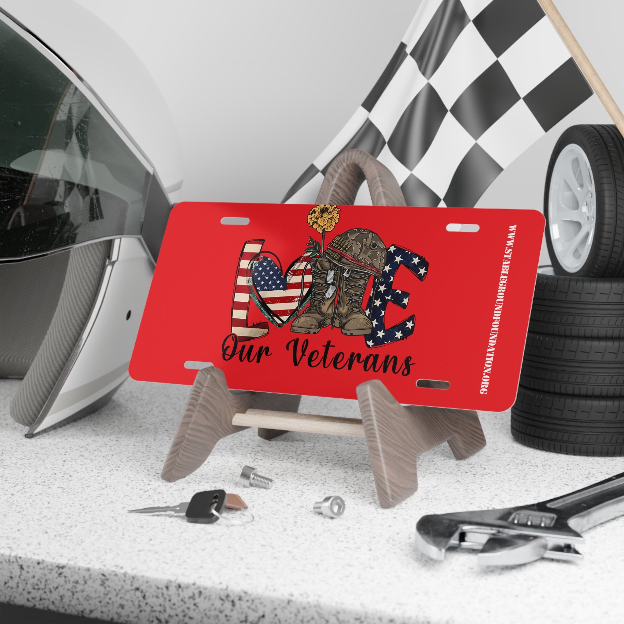 Red license plate featuring the word “LOVE” with an American flag heart, camouflage combat boots holding a yellow flower, and patriotic lettering. Text below reads “Our Veterans.”