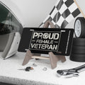 Black license plate with distressed white bold text reading: “Proud Female Veteran” inside a rectangular frame. Displayed on a wooden stand with tools, keys, helmet, and checkered flag in the background.