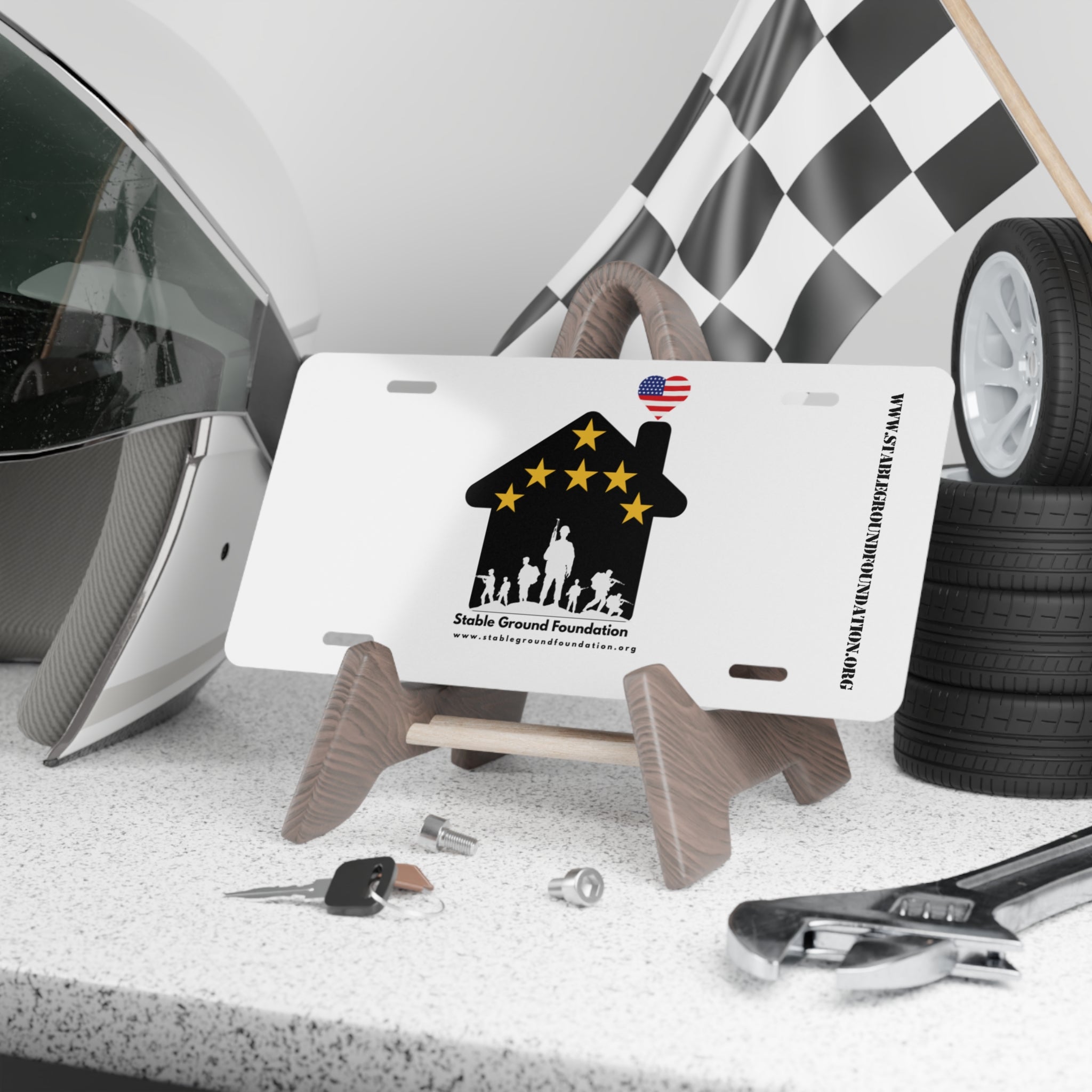 White license plate featuring Stable Ground Foundation logo with a black house, gold stars, military family silhouettes, and an American flag heart.